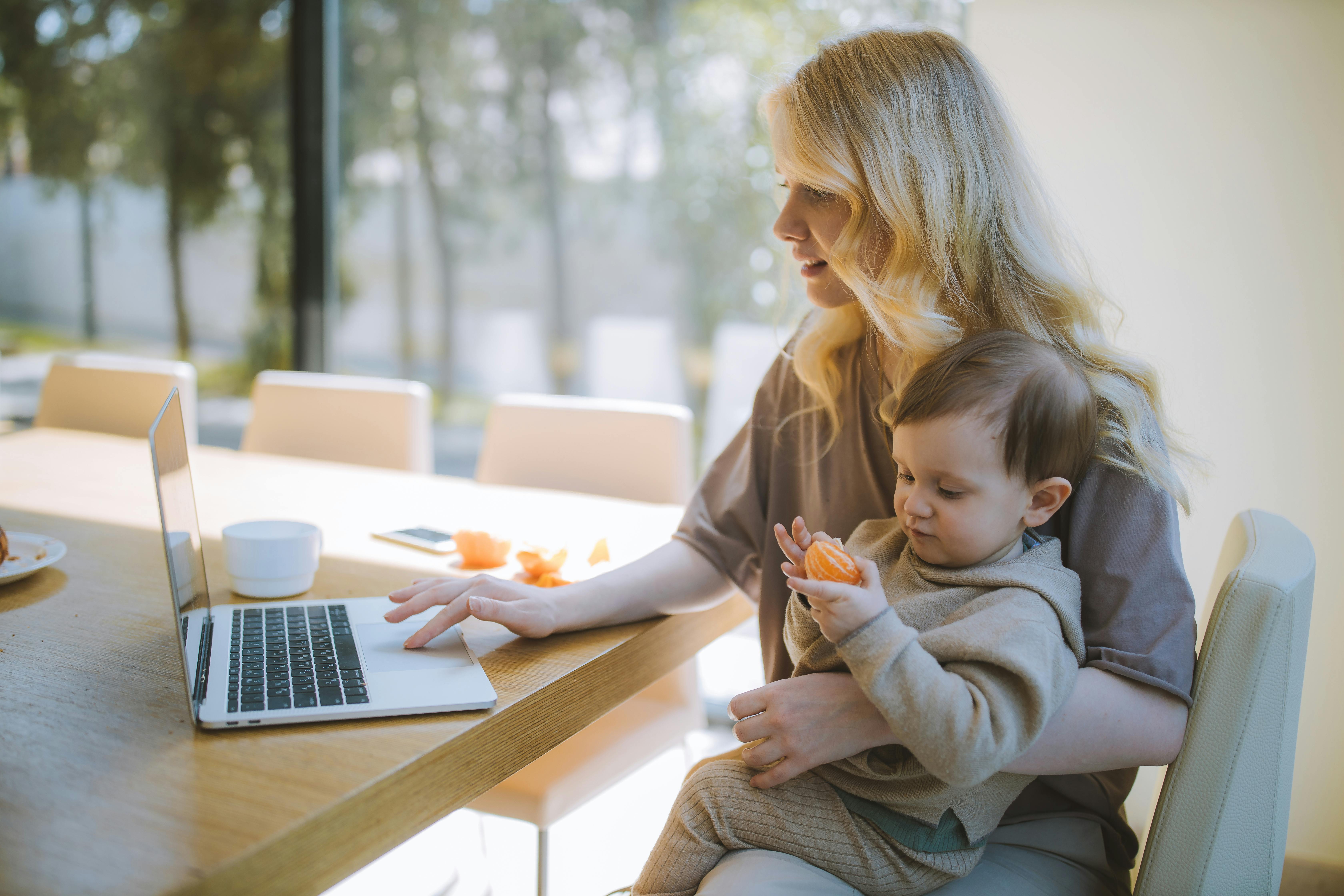 Mother working on a laptop at a sunlit kitchen table with her toddler on her lap
